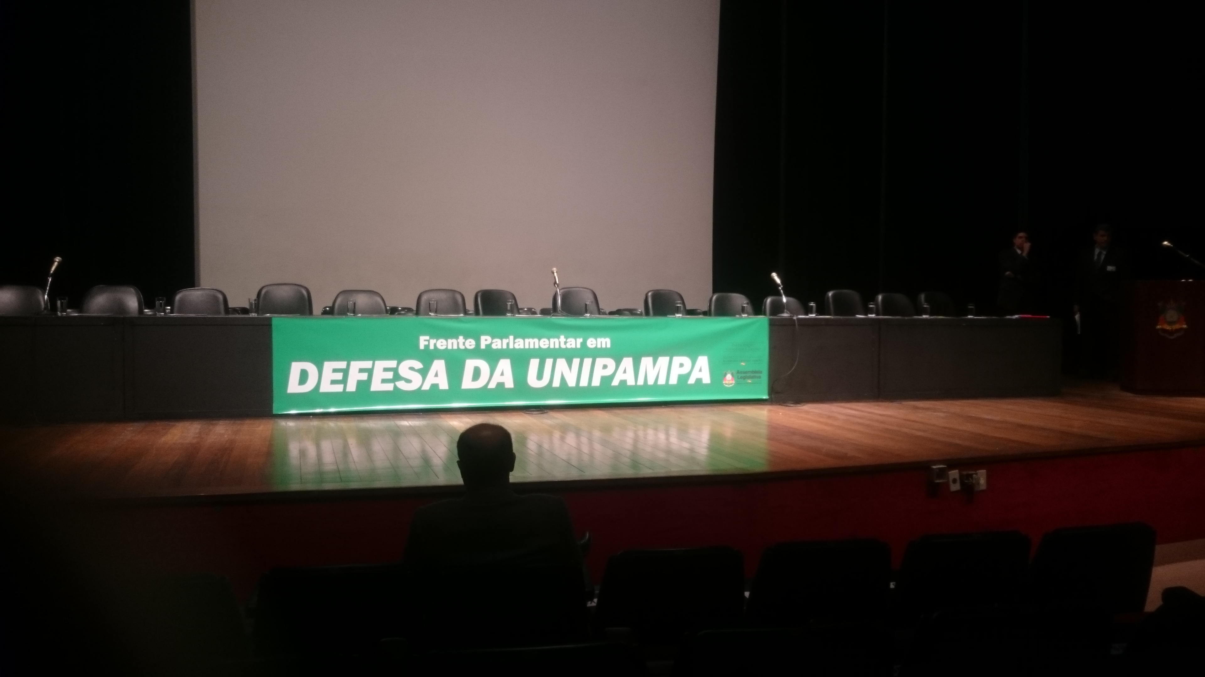 Palco da Assembleia Legislativa de Porto Alegre sem pessoas e com uma faixa verde: Frente Parlamentar em Defesa da UNIPAMPA
