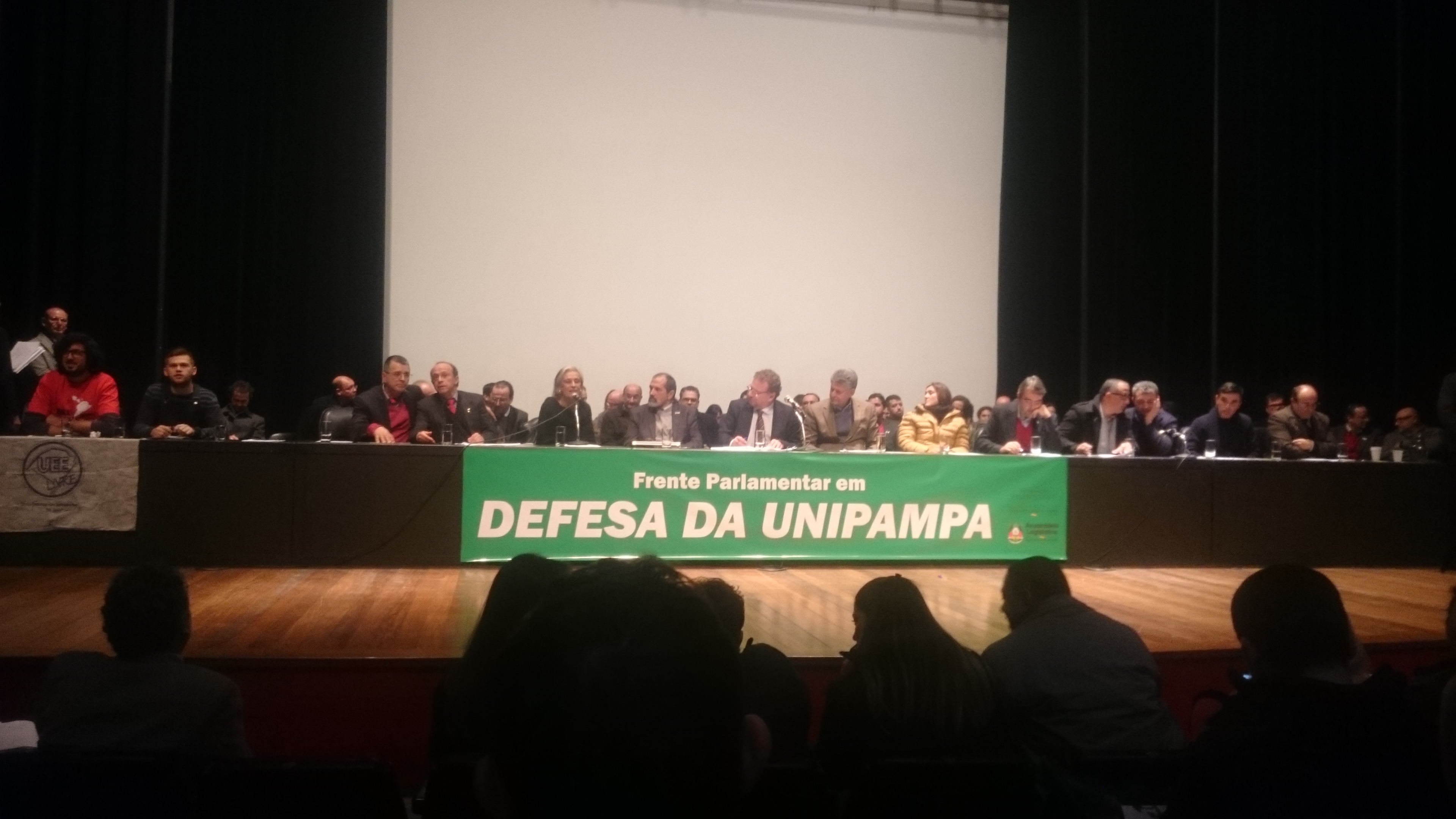 Palco da Assembleia Legislativa de Porto Alegre com a mesa de representantes parlamentares e da UNIPAMPA e com uma faixa verde: Frente Parlamentar em Defesa da UNIPAMPA